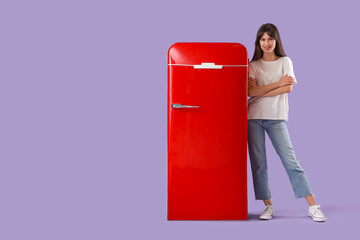 Beautiful young happy woman near red fridge on purple background