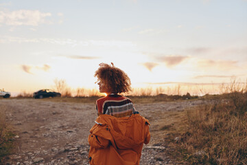 Fototapeta premium Woman with curly hair wearing rainbow sweater and orange jacket outdoors, smiling, holding smartphone, autumn landscape, warm sunlight, casual lifestyle, relaxed mood.