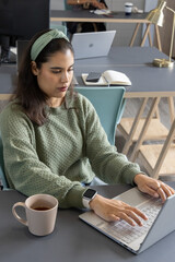 Asian woman in green sweater working at desk in workspace with silver laptop, pink mug, smartwatch