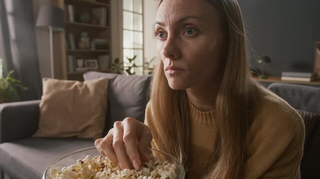 Medium close-up of face of young Caucasian woman watching horror or thriller film on TV while snacking on bowl of popcorn, jumping with fear, displaying genuine emotions of shock and dread