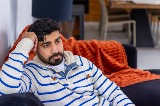 Asian adult male reclining on sofa in living room with orange throw, wearing striped knit hoodie