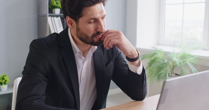Thoughtful businessman in suit sitting at desk, looking at laptop screen during work in office. Pensive executive thinking about difficult work task, analyzing information or making serious decision.