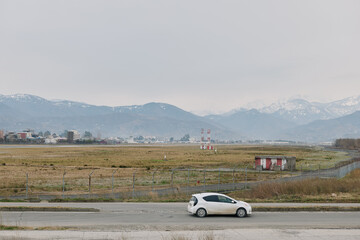 Fototapeta premium White compact car parked on roadside near field with distant mountains under cloudy sky. Concept of travel, transportation, countryside, and nature landscape in calm weather conditions.