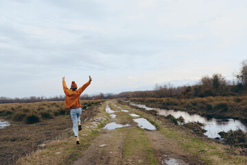 Fototapeta premium A woman lifestyle concept showing a carefree female walking on a muddy rural path with arms raised, wearing casual clothes in an open natural landscape under a cloudy sky.