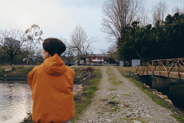 Fototapeta premium A woman in an orange jacket enjoys a peaceful outdoor lifestyle by a lake, standing on a gravel path near a wooden bridge surrounded by trees and nature.