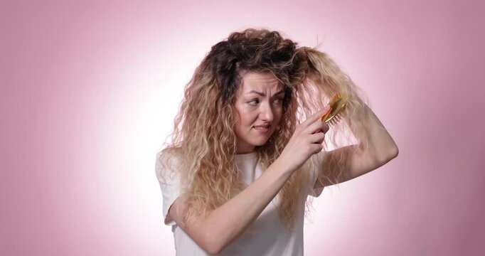 Unhappy woman trying to brush her tangled hair on pink background