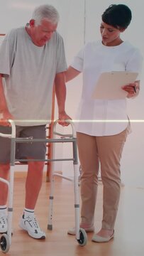 Helping therapist aiding senior man wearing gray tee taking steps in clinic with walker, clipboard