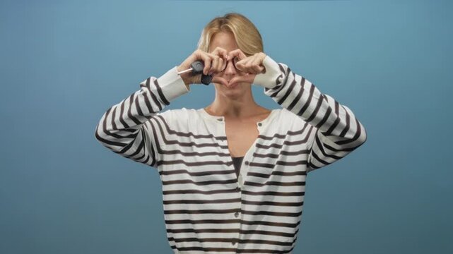 Woman forms a heart shape with hands over face while holding car keys and wearing a striped cardigan in studio; playful.