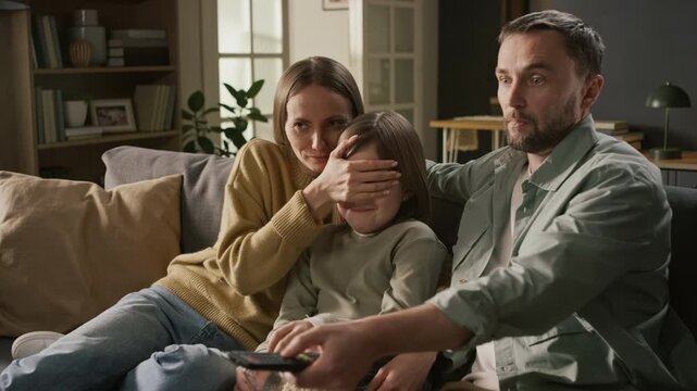 Medium shot of sensitive young Caucasian couple watching TV with daughter, protecting child from explicit content by covering eyes, then switching off movie with remote control