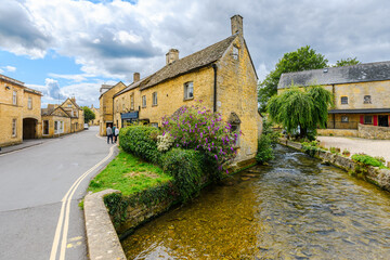 Fototapeta premium The shallow River Windrush passes warm toned Cotswold stone buildings in the center of the village of Bourton-on-the-Water, England, in the Cotswold district.
