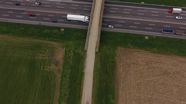 Urban highway intersection: An elevated pedestrian walkway arches over a bustling highway, where various vehicles traverse its lanes, while a lush green field and a harvested field border the scene