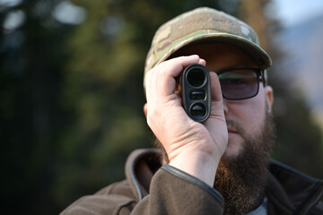 Fototapeta premium Bearded man using a rangefinder while hunting in the outdoors with camouflage cap and glasses