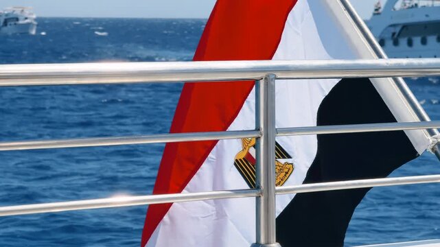 Close up of yacht deck fenced safety stainless railing and Egyptian national flag behind. Excursion boats floating in the background