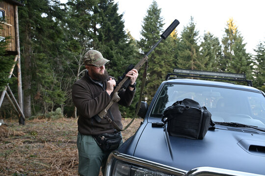 Hunter preparing a scoped rifle beside an off-road vehicle in a forested hunting camp
