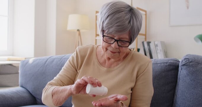 Senior woman taking pills to cure disease, taking medicaments regularly, painkillers to relieve pain, sitting on comfortable sofa at home. Old people, pharmacy and treatment concept