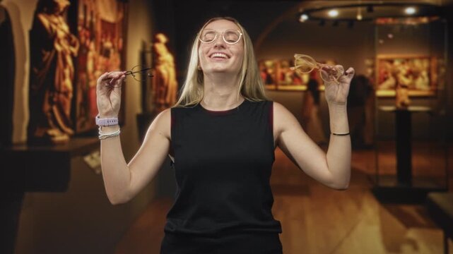 Woman holding two pairs of glasses, smiling and holding them up by each hand while testing eyewear in a museum building with paintings and sculpture visible; joyful curiosity.