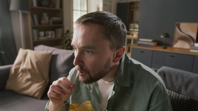 Medium close-up of face of young Caucasian man sitting alone on couch at home at leisure time, watching horror or thriller movie on TV, eating chips, with emotions of fear, dread, astonishment, dismay