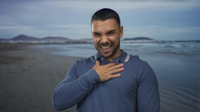 Young hispanic man with hand on chest standing on sandy beach shore by calm ocean water waves; serenity.