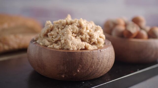 Freshly ground hazelnut butter in small wooden bowl with whole nuts and bread in background