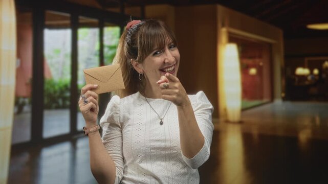 Woman holding envelope and pointing finger in hotel building lobby while smiling and looking at camera; playful invitation.
