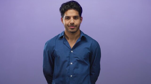 Young man standing with hands behind back in studio, wearing a blue button shirt against a purple backdrop, eyes moving from closed to open with a slight smile; calm confidence.