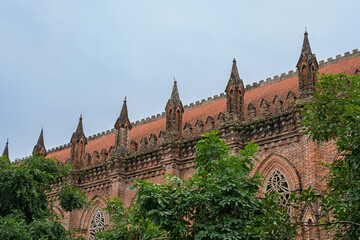 Fototapeta premium Gothic brick facade of Chau Son Monastery with decorative arches and pointed spires partially covered by lush greenery under overcast sky.