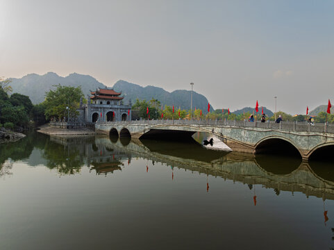 Curved stone bridge with red flags crossing tranquil water toward ancient temple gate in Hoa Lu with limestone mountains in background.