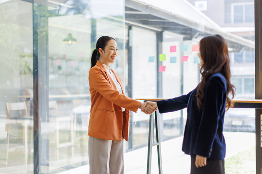 A scene depicting two women in blazers engaging in a friendly handshake in a well-lit office, illustrating the importance of greeting and goodwill in business interactions.