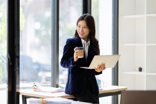 A young professional woman stands confidently with a laptop and a coffee mug in a modern office, representing the essence of work-life balance and productivity.