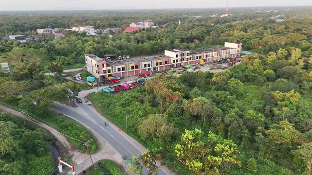 The cinematic drone shot moves forward toward a row of colorful shop houses along a rural road, surrounded by lush tropical vegetation under the warm sunlight.