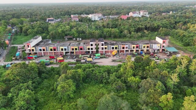 An aerial shot showing a row of shop houses surrounded by tropical forest, with various trucks parked in front of them.