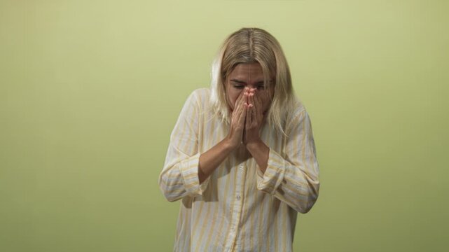 Woman young blonde hispanic covering hands over face, pressing palms to nose and mouth, wearing yellow striped shirt against pale green studio wall; discomfort illness.