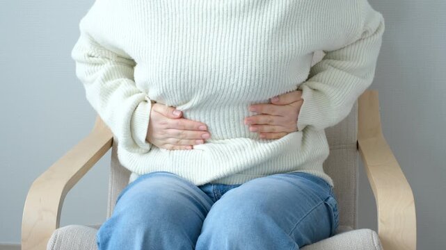 Young woman sitting on chair and holding her stomach with her hands, abdominal pain concept.
