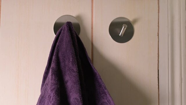 A man hangs two purple towels on metal hooks on the wall. Male hands alternately hang clean purple towels on round self-adhesive hooks fixed to light tiles in the bathroom
