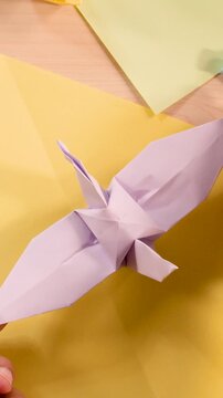 Hands Flapping Purple Origami Bird Wings Over Yellow Paper Background