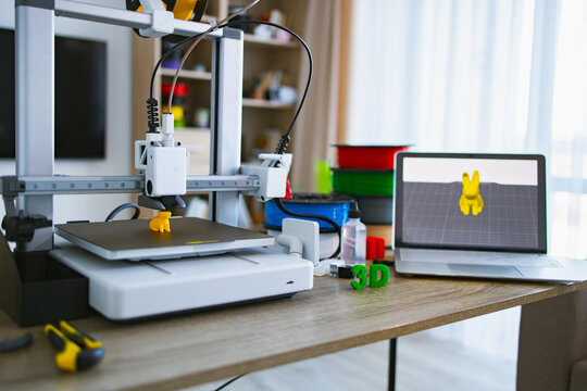 A 3D printer is actively creating a small yellow object on its build plate, with filament spools and a laptop displaying the design nearby