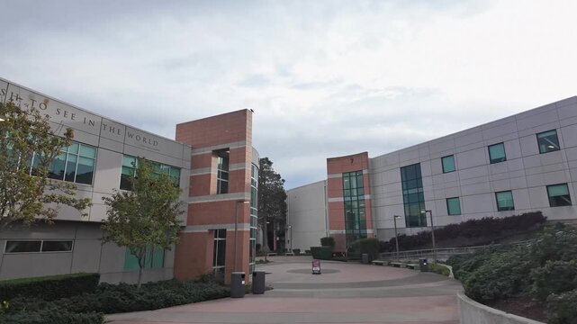 Wide shot walking through the modern architectural grounds of Skyline College campus in San Bruno, California, on a cloudy day, featuring a quote by Gandhi inscribed on the building facade