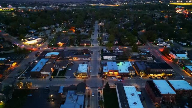 An aerial perspective of a quiet small town intersection at dusk, featuring glowing storefronts, streetlights, and sparse traffic in a suburban setting. Oswego, Illinois.