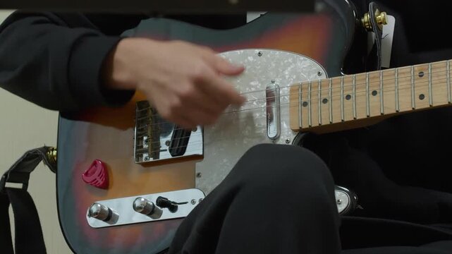 Close-up shot of a musician's hand strumming a sunburst Telecaster-style electric guitar. The guitarist plays a riff, showing skill and passion in a studio or home setting. Dynamic and focused.