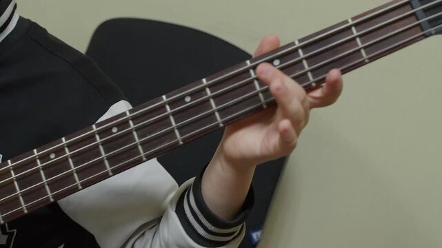 Close-up video of a child's hands playing a black electric bass guitar. The camera pans from the plucking hand up the fretboard to the fretting hand. Focus on musical practice and learning.