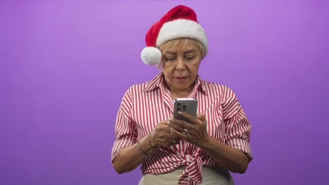 Woman in santa hat taps smartphone with hands against purple studio backdrop wearing striped shirt and bracelets; quiet contemplation.