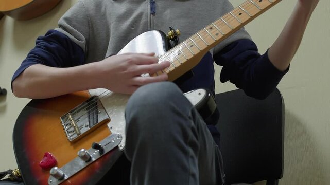 A young man or teenager sits and plays a sunburst electric guitar. Close-up shot focuses on his hands moving quickly over the fretboard during a practice session or rehearsal.