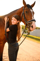 Obraz premium Smiling girl hugs horse while standing outside at sunset, looking happily at camera. Tender bond between rider and horse. Horse care concept.