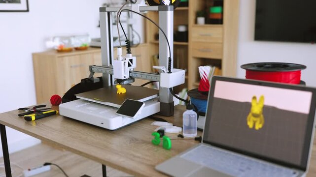 A 3D printer is actively creating a small yellow toy figure on its build plate, with filament spools and tools nearby