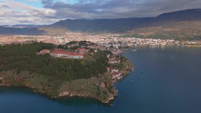 A wide panoramic sweep across Lake Ohrid, approaching the historic old town center before tilting down for a majestic reveal of St. John at Kaneo church perched high above the crystal-clear water.