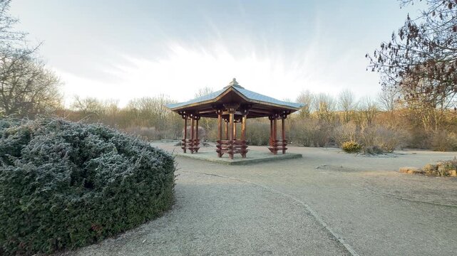 A beautiful wooden pavilion stands in a quiet clearing surrounded by frost-covered bushes. The soft morning sky creates a feeling of peaceful isolation in the crisp winter air.