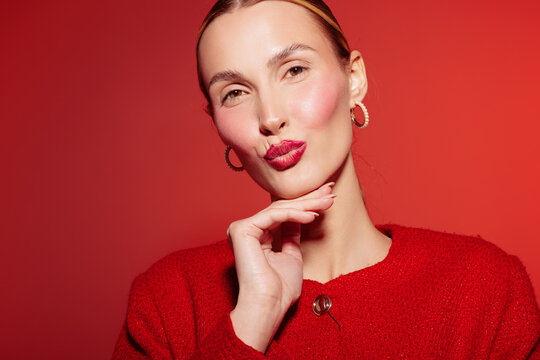 Young woman with red lipstick and earrings poses with hand near chin on red background. Fashion portrait with confident expression and stylish makeup in studio lighting.
