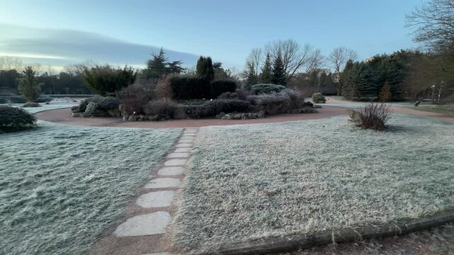 A line of flat stones crosses a frost-covered lawn toward a circular garden bed. Evergreen trees and bushes stand still under a pale morning sky. The atmosphere is crisp, organized, and serene.