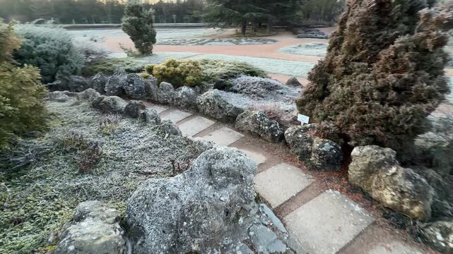 A winding stone path leads through a rugged rock garden covered in morning frost. The quiet morning light creates a peaceful and refreshing mood across the park. The air feels crisp and still.