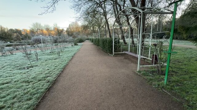 Elegant white metal arches stand over a quiet bench along a gravel walkway. The frost-covered grass and dormant bushes create a cold, nostalgic atmosphere in this peaceful park setting.